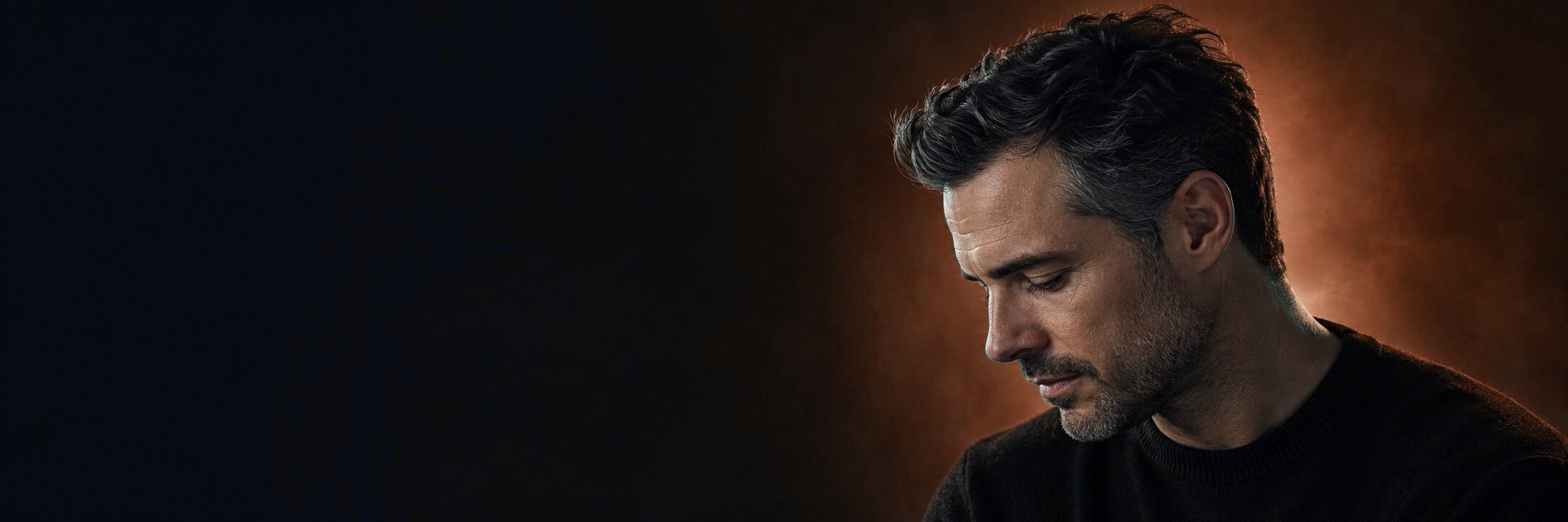 A man with dark, swept-back hair, looking down